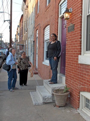 A woman is standing in front of her door on top of a set of 3 white marble steps, smiling at the camera.  Nanta and Dona are on the sidewalk, and Dona is talking to the woman.  Along the wall, about 10 feet away is another door, with only one short step in front of it, and the wall extends for the rest of the block, with rows of houses all connected together ('row houses')